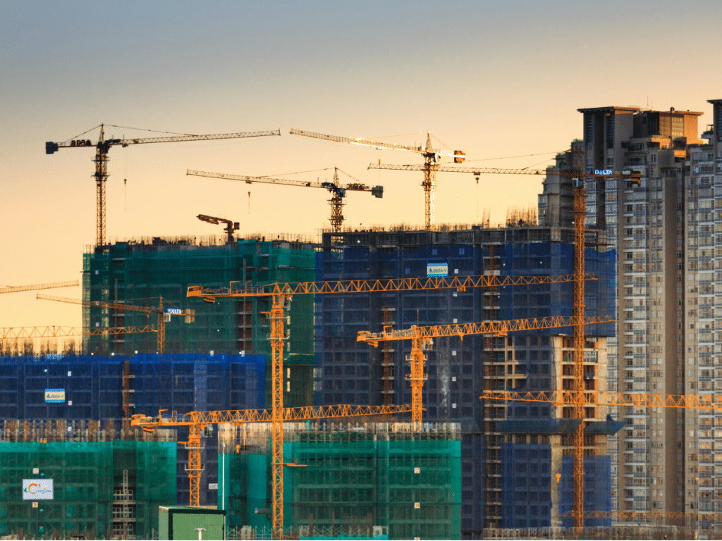 Busy construction site with multiple cranes next to buildings under construction, many covered with blue and orange safety netting.